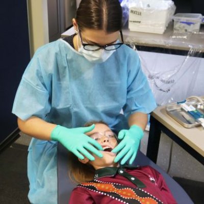 A woman in a blue gown, green gloves and facemask looks at the teeth of a child lying in front of her